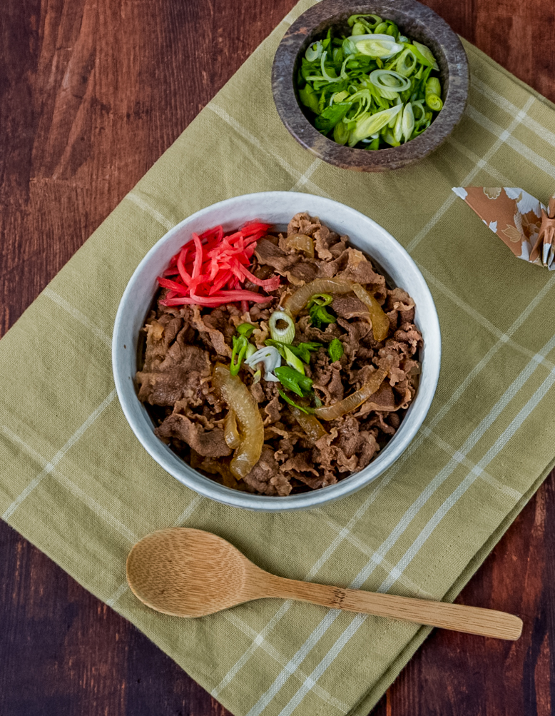 Eine Schüssel Gyudon (japanische Reis-Bowl mit Rindfleisch und Zwiebeln) serviert mit Frühlingszwiebeln und Beni Shouga (eingelegtem Ingwer) auf einer gelben Stoffserviette, daneben ein Holzlöffel.