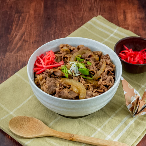 Eine Schüssel Gyudon (japanische Reis-Bowl mit Rindfleisch und Zwiebeln) serviert mit Frühlingszwiebeln und Beni Shouga (eingelegtem Ingwer) auf einer gelben Stoffserviette, daneben ein Holzlöffel.